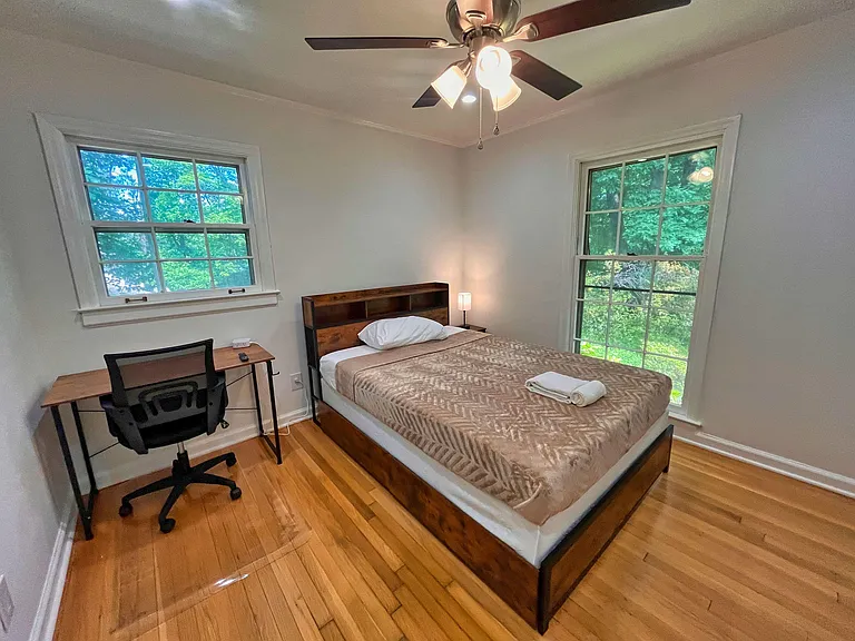 Furnished bedroom with hardwood floors, a work desk, ceiling fan, and large windows overlooking greenery.