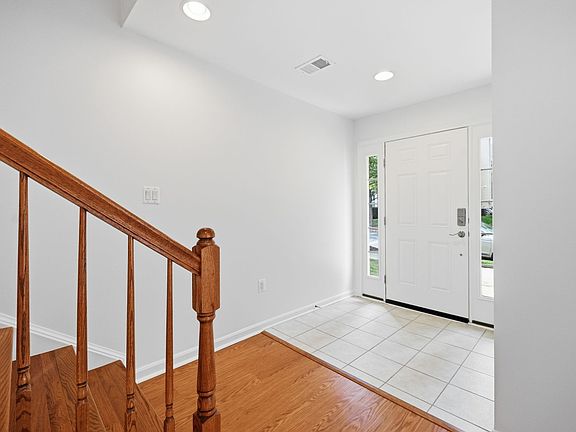 A bright and welcoming entry featuring easy-care tile flooring, a side-lit front door for natural light, crisp neutral walls, recessed lights, and a hardwood staircase.