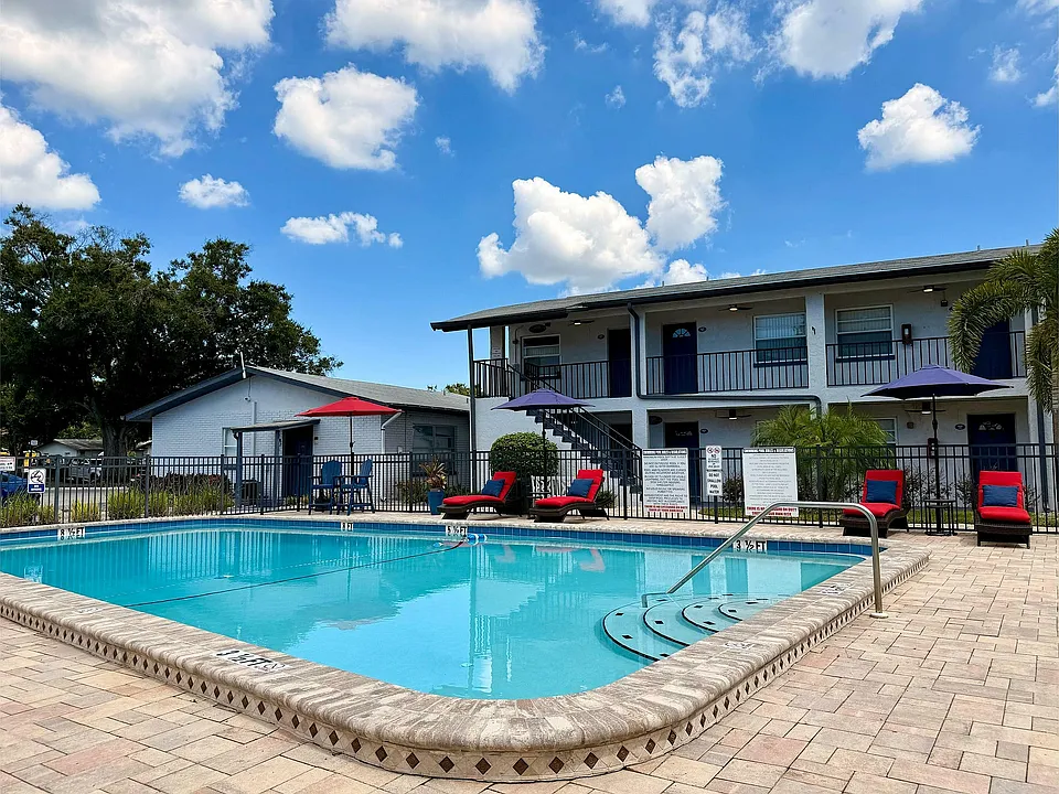 Charming apartment complex with a sparkling pool surrounded by sun loungers, vibrant umbrellas, and lush greenery under a bright blue sky.