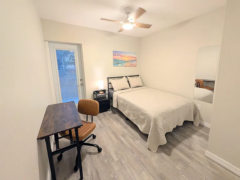 Furnished bedroom with a full bed, desk workspace, ceiling fan, and direct outdoor access through a glass door.