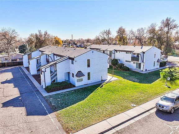 Charming apartment complex with white exteriors, lush green lawns, and ample parking in a serene neighborhood setting.
