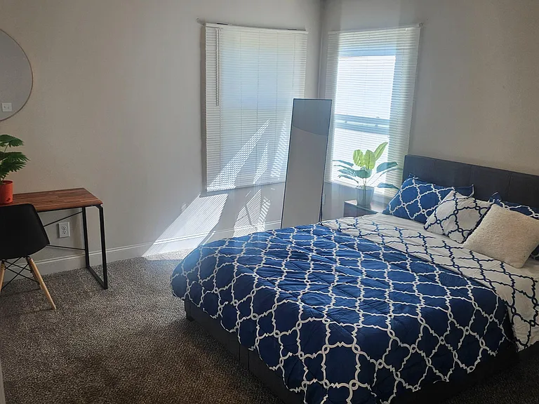 Carpeted bedroom with a full bed, work desk, and two windows with blinds for comfortable daily living.