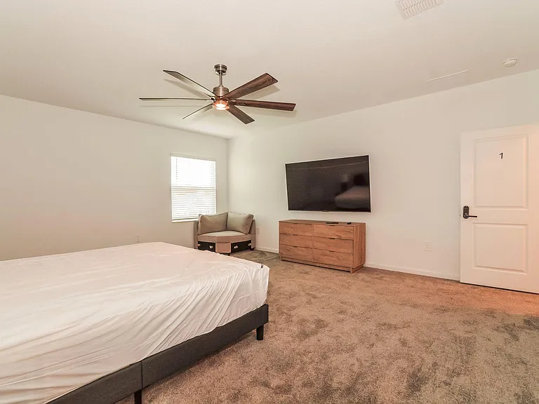 Spacious bedroom with carpet flooring, ceiling fan, window with blinds, and a mounted TV with dresser for storage.