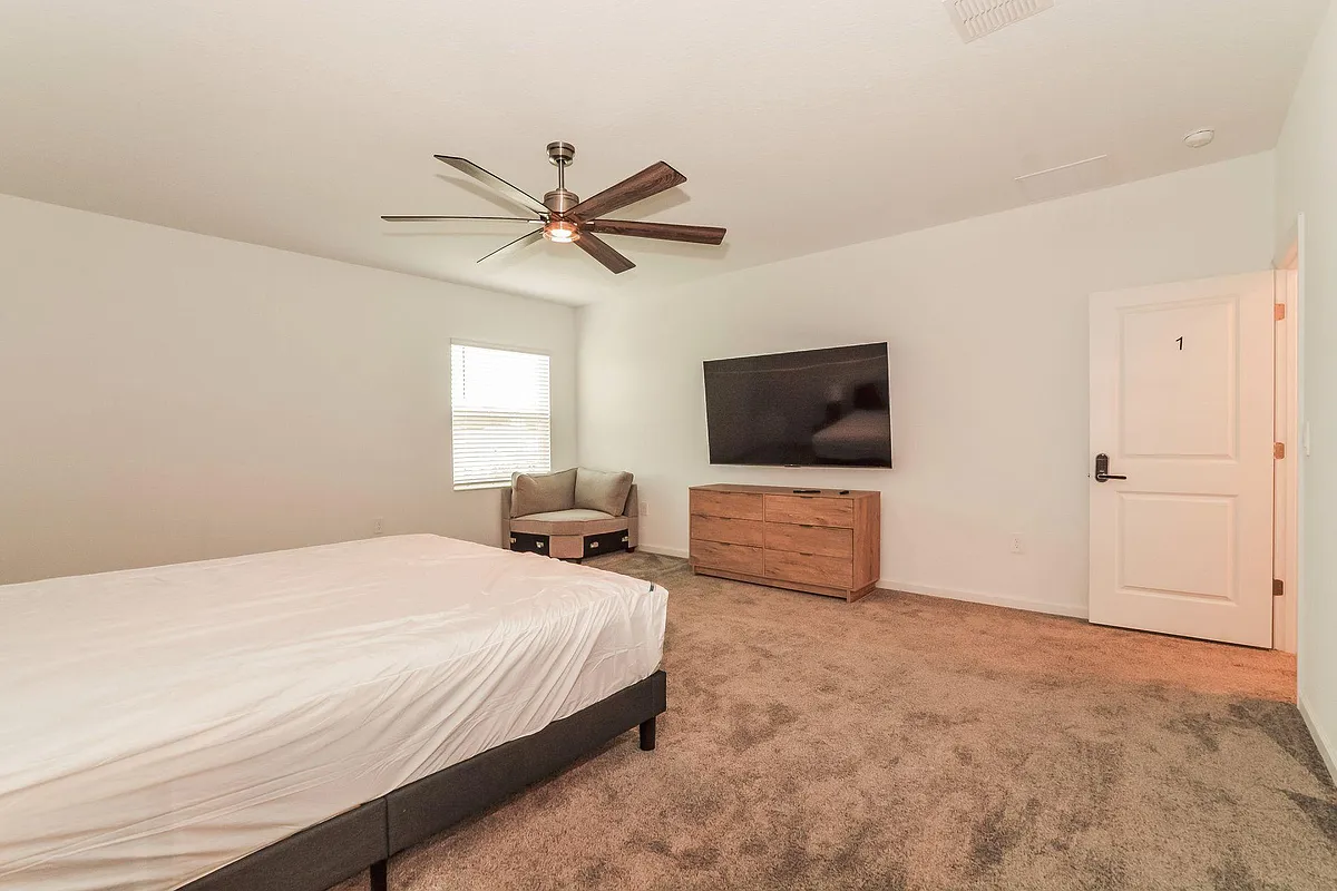 Spacious bedroom with carpet flooring, ceiling fan, window with blinds, and a mounted TV with dresser for storage. - Slash Way