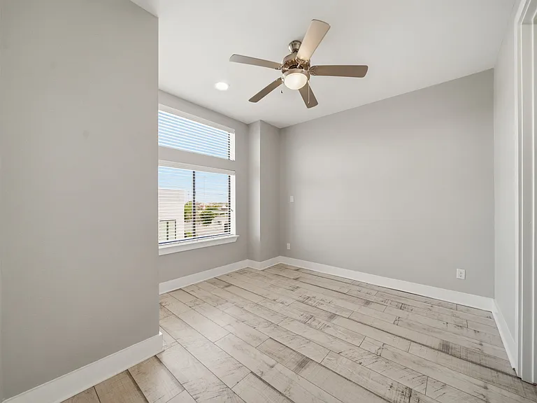 Bright, modern third-floor bedroom featuring light wood flooring and neutral gray tones throughout. Large windows fill the space with natural light, while a ceiling fan adds comfort and functionality.