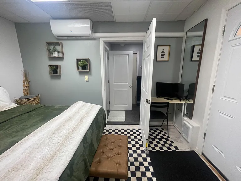 Furnished bedroom with a bed and bench, wall decor, and a dedicated desk setup with monitor and full-length mirror.
