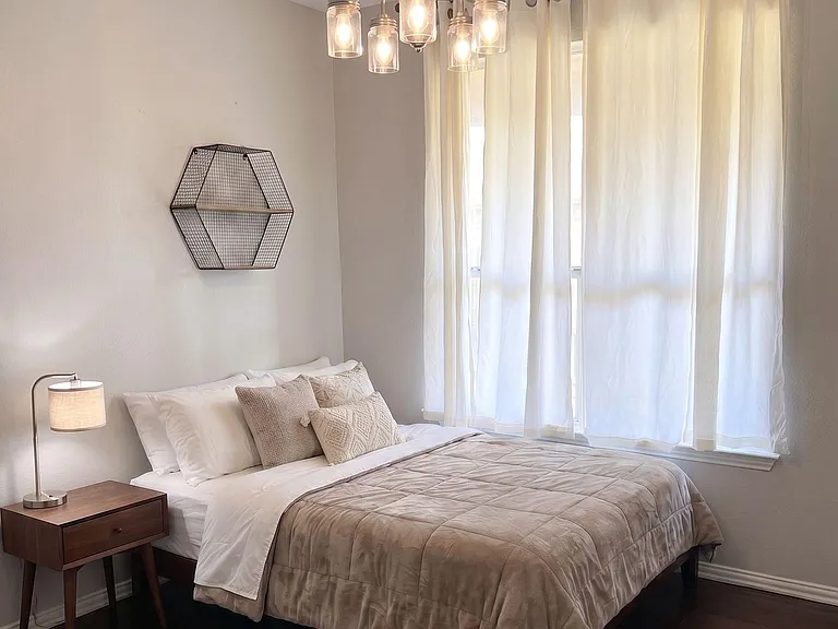 Well-sized bedroom with a comfortable bed, bedside table, and large window with curtains plus an overhead light fixture.