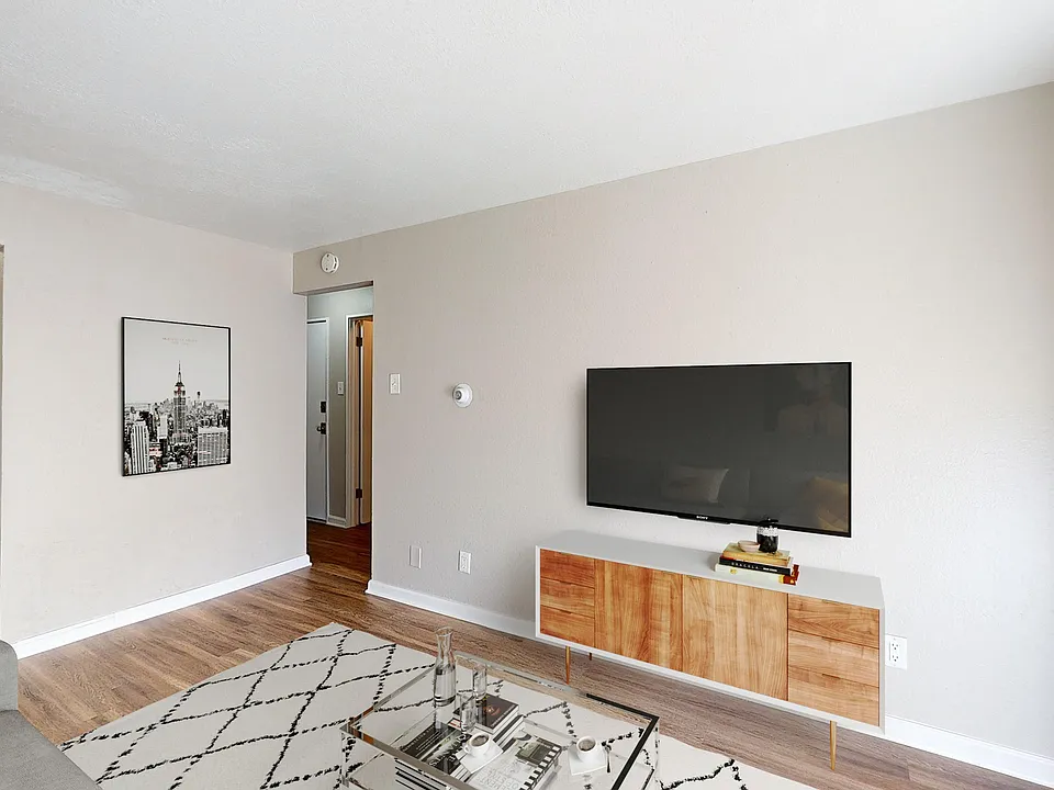 Modern living room with natural light, sleek wooden flooring, and a stylish TV unit. Adjacent kitchen area with classic cabinetry visible.