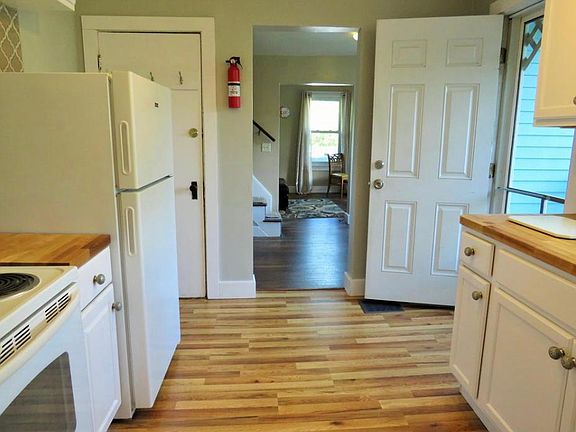 View from bathroom doorway of kitchen, leading to dining room and living room beyond. Door behind fridge is a storage/pantry area.