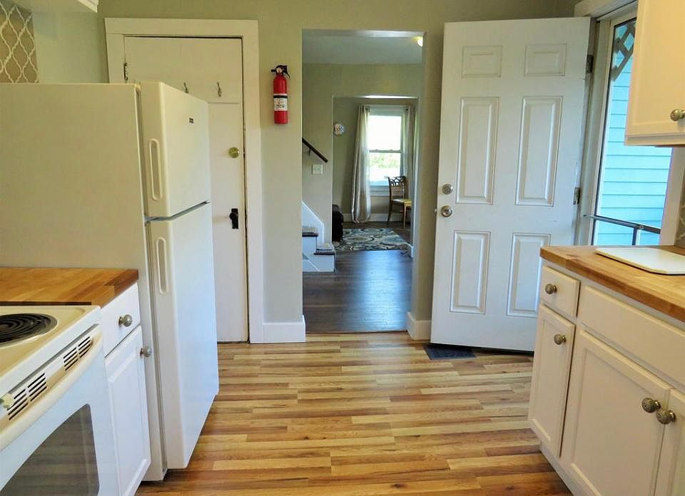 View from bathroom doorway of kitchen, leading to dining room and living room beyond. Door behind fridge is a storage/pantry area.