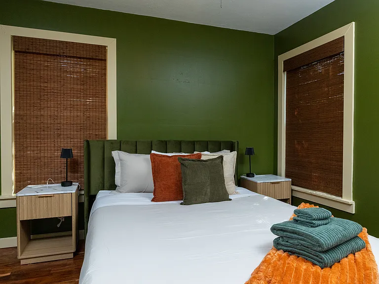 Relax in this serene bedroom featuring rich green walls, warm wood floors, and a plush bed with cozy accent pillows. A ceiling fan, natural textures, and soft lighting create a peaceful, modern retreat.