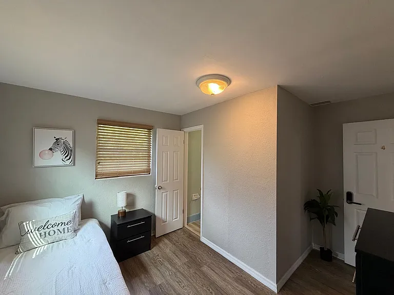 Furnished bedroom with a comfortable bed, nightstand, and window blinds, plus easy access to the hall and adjacent bath.