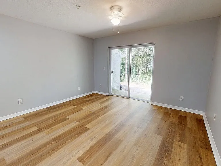 Bright, empty bedroom with sleek hardwood floors and a sliding glass door leading to a lush outdoor view.