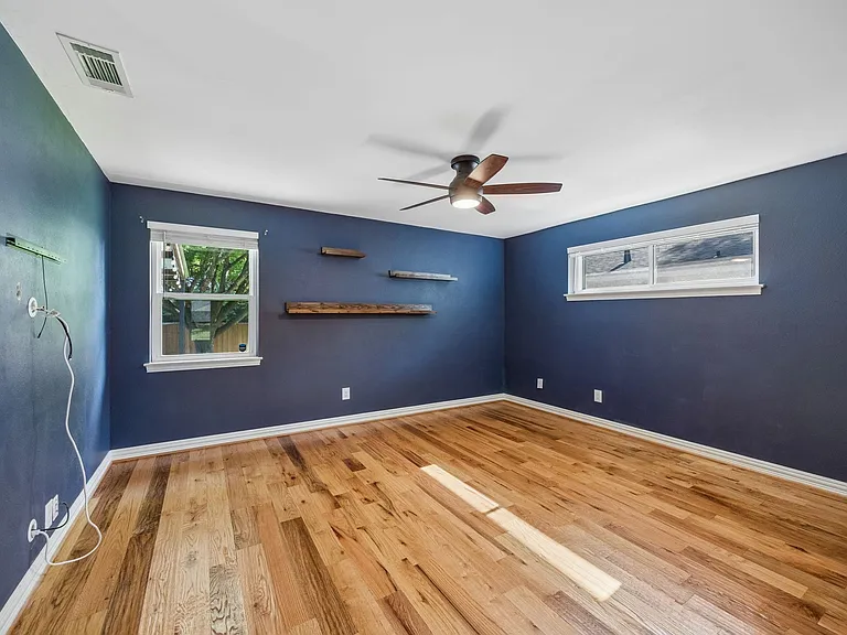 Spacious room featuring wood-finish flooring, a ceiling fan with integrated lighting, two windows with white trim, and built-in floating shelves