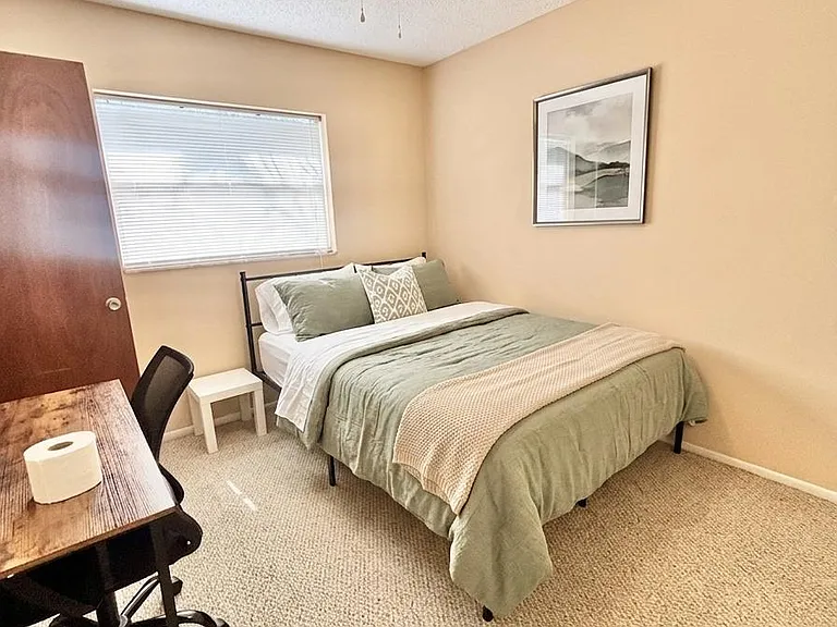 Private bedroom with a furnished bed, nightstand, and dedicated desk workspace, plus a window with blinds and ceiling fan.