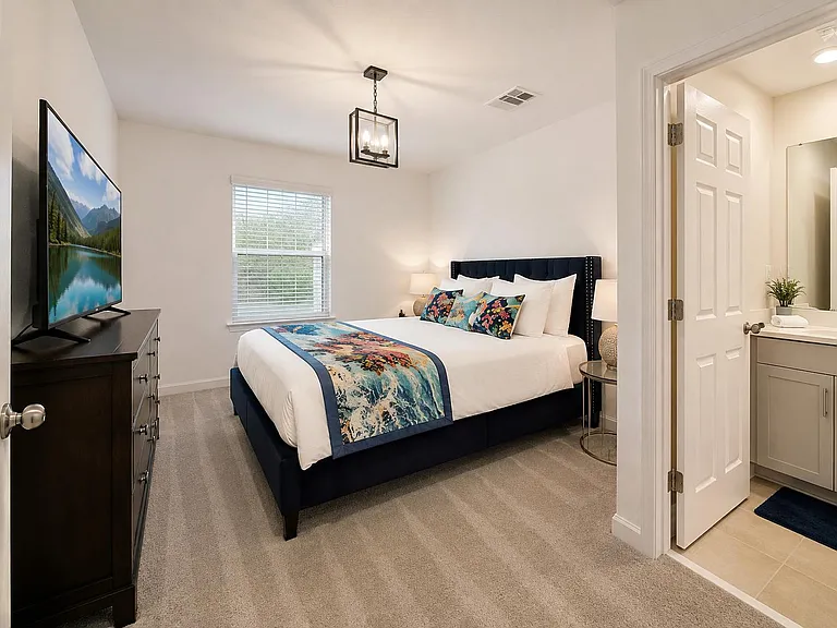 Spacious bedroom with a large window, plush carpeting, dresser/TV setup, and direct access to a private vanity area.