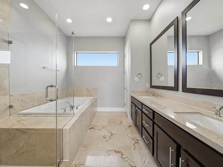 Luxurious primary bathroom showcasing a generous layout with dual vanities, sleek modern countertops, and refined marble tile finishes. A deep soaking tub and separate glass-enclosed shower create a spa-like retreat, enhanced by natural light.