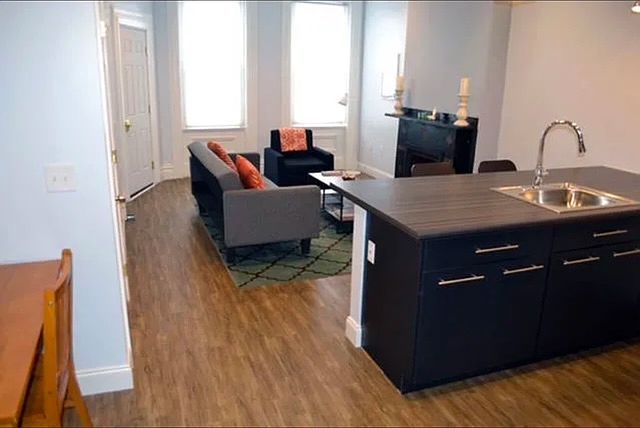 View from the kitchen, looking back at the private entrance door to the left, and 2 tall front windows lighting up the spacious room.