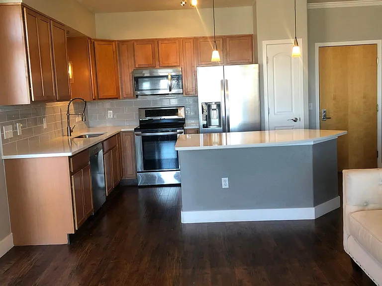 View of the kitchen from the living room: this spacious unit has a large island with overhanging countertop to allow for barstools. Multiple luxury finishes have been added, such as luxury marble-look quartz countertops and stainless steel appliances