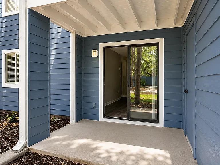 Inviting apartment entryway featuring elegant blue siding and a modern glass door opening to a serene garden view.