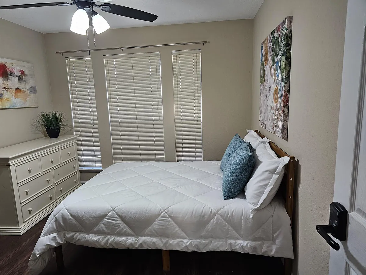 Comfortable bedroom with a furnished bed, dresser storage, ceiling fan, and three tall windows with blinds. - Sabinal Trl