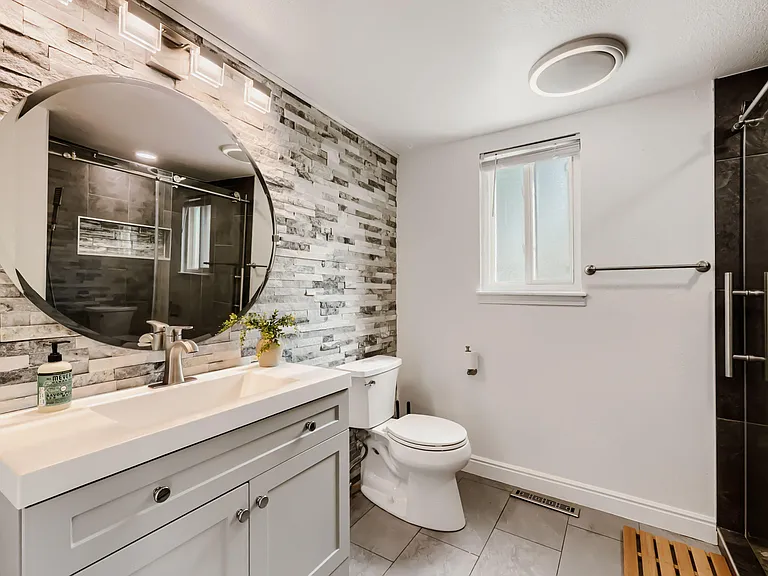 Updated bathroom with single-sink vanity, round mirror, tiled flooring, and a glass-enclosed shower plus a frosted window.