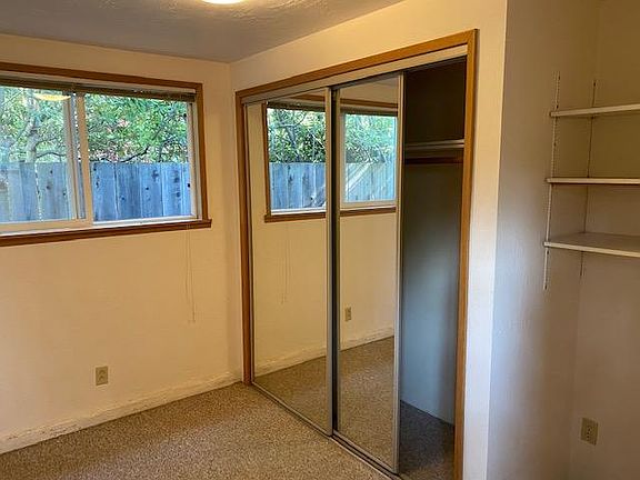 Small carpeted bedroom with full sliding mirrored door closet and nook for dresser or desk.