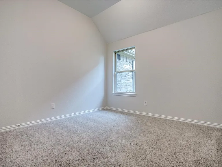 Carpeted room with a vaulted ceiling, light-colored walls, and a single window offering a view of exterior brickwork