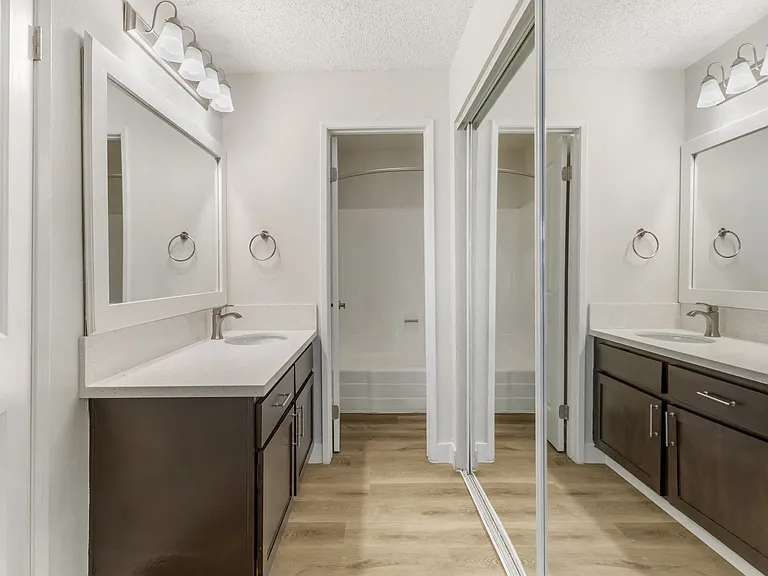 This modern bathroom features wide vanities with sleek quartz countertops, framed mirrors, and stylish lighting. A mirrored closet and separate shower area complete the bright, functional space.