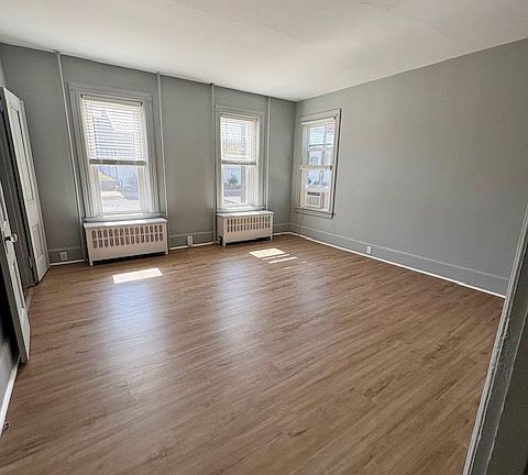 Three tall windows line one side of the room, allowing ample natural light to pour in. Each window is fitted with white blinds and sits above classic white radiators. The ceiling is white, and the room has clean, minimalistic!