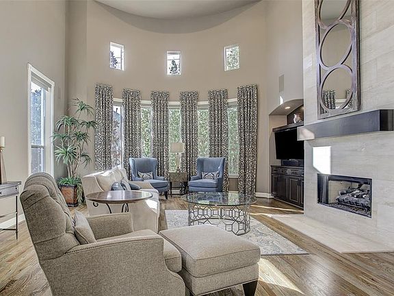The two-story living room is bright and spacious with a gorgeous curved wall of windows, dramatic natural stone fireplace, and gleaming hardwood floors.