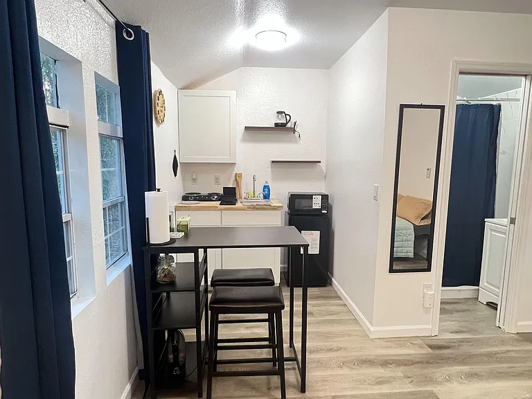 Efficient kitchenette with open shelving, compact appliances, and a bar-height dining setup beside large windows.