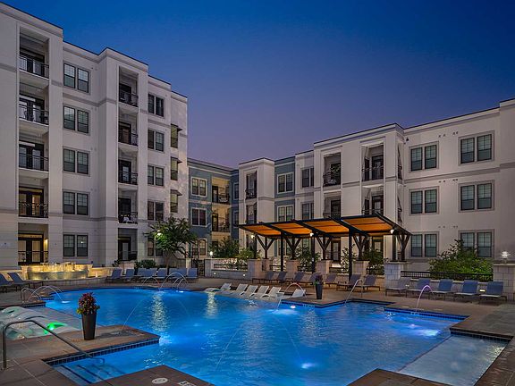 Dusk shot of pool with in-water lounge seats, additional lounge chairs, lighted fountain and covered area