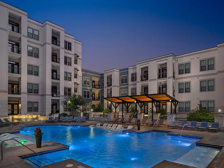 Dusk shot of pool with in-water lounge seats, additional lounge chairs, lighted fountain and covered area