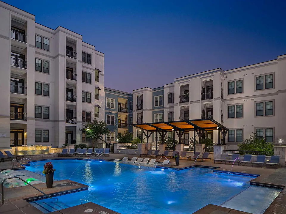Dusk shot of pool with in-water lounge seats, additional lounge chairs, lighted fountain and covered area