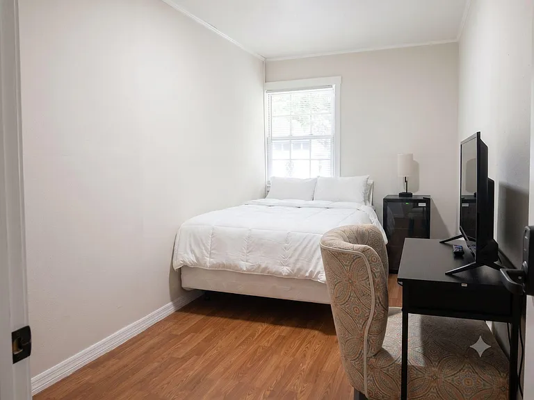 Furnished bedroom with a comfortable bed, side table and lamp, plus a dedicated desk setup and window with blinds.