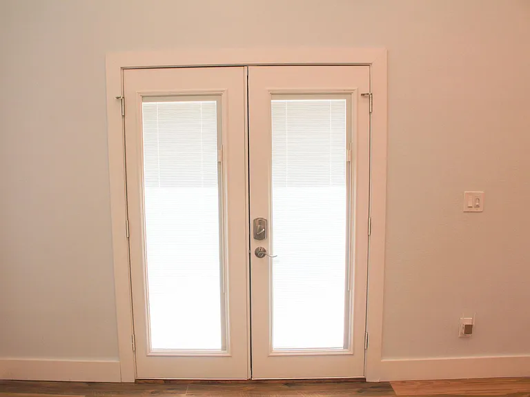 View of double-door with built-in blinds closed with keyless deadbolt entry. Light switch and separate ceiling fan control at entrance.