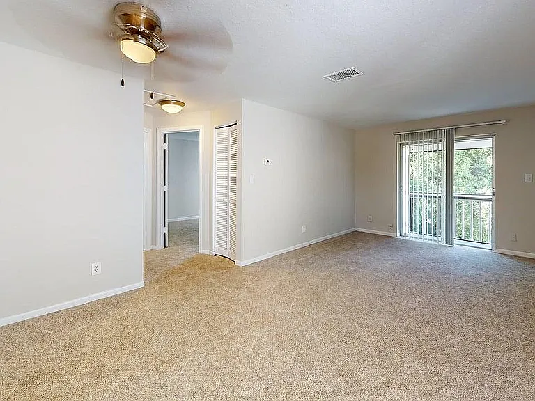 Bright and spacious living room with large windows, plush carpeting, and modern ceiling fan, perfect for comfortable living.
