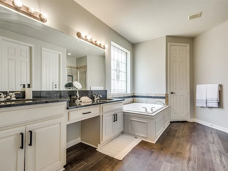 Bathroom featuring dual vanities with dark countertops, built-in soaking tub with decorative tile surround, a leaded glass window, and wood-finish flooring
