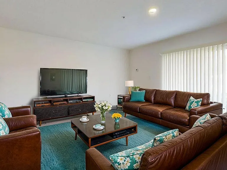 Inviting living room with elegant brown leather sofas, a sleek flat-screen TV, and a vibrant teal rug accented by floral pillows and fresh flowers.
