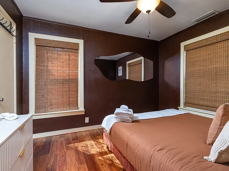 Cozy retreat with warm tones and natural light. This comfortable bedroom features a queen bed, bamboo shades, a ceiling fan, and a modern mirror accent _ perfect for unwinding after a day exploring Dallas.
