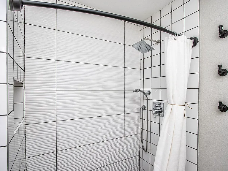 Modern bathroom shower at Casa Del Sol, Tempe furnished rental. Features sleek white grid and wavy textured tile, a luxury rain showerhead, a handheld sprayer, industrial black pipe hooks, and an inset storage niche. Premium amenity for a Tempe, AZ stay.