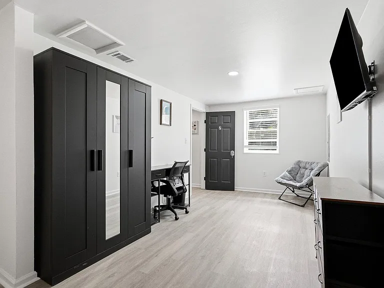 Furnished bedroom with large wardrobe, dedicated desk, wall-mounted TV, and a window for natural light.