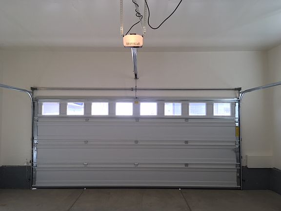 View of oversized two-car garage. Interior has been painted and a motion-activated overhead light has been installed. A faux wood window covering covers the window. 2 extra-large recycle bins and 1 extra-large regular trash bin are provided.