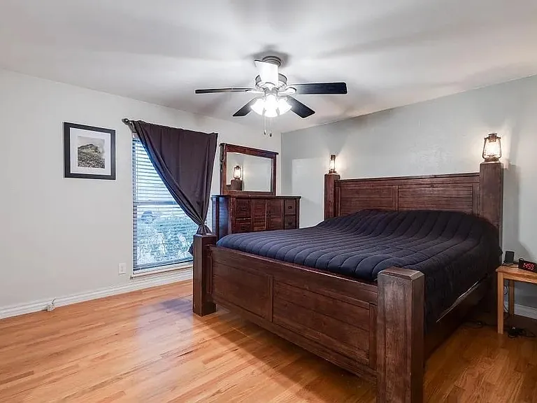Spacious bedroom featuring wood-finish flooring, a ceiling fan with integrated lighting, window with blinds, and light-toned walls