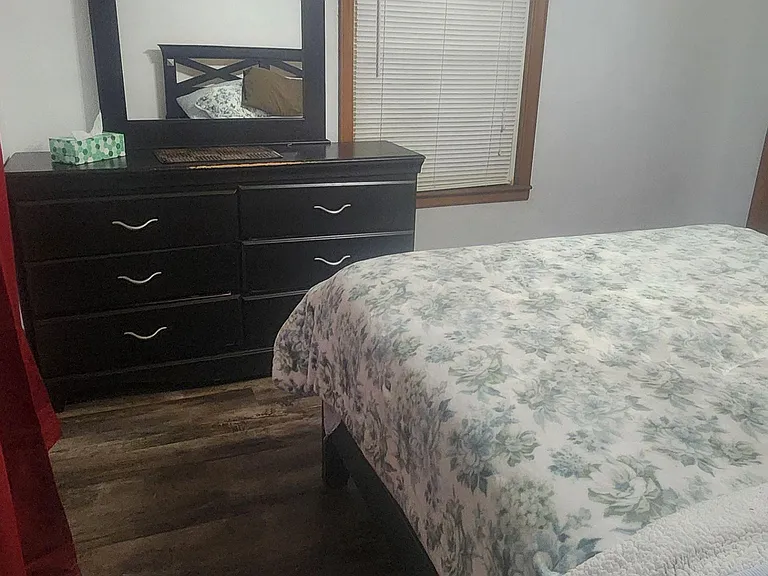 Furnished bedroom featuring a comfortable bed, dresser with mirror, and a window with blinds for added privacy.