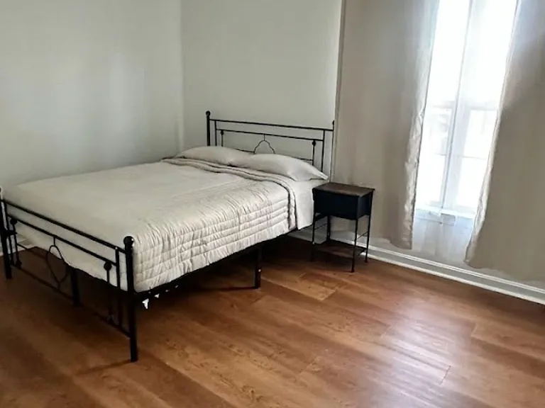 Furnished bedroom with a full-size bed, nightstand, and hardwood-style flooring, plus a window with curtains for daylight.