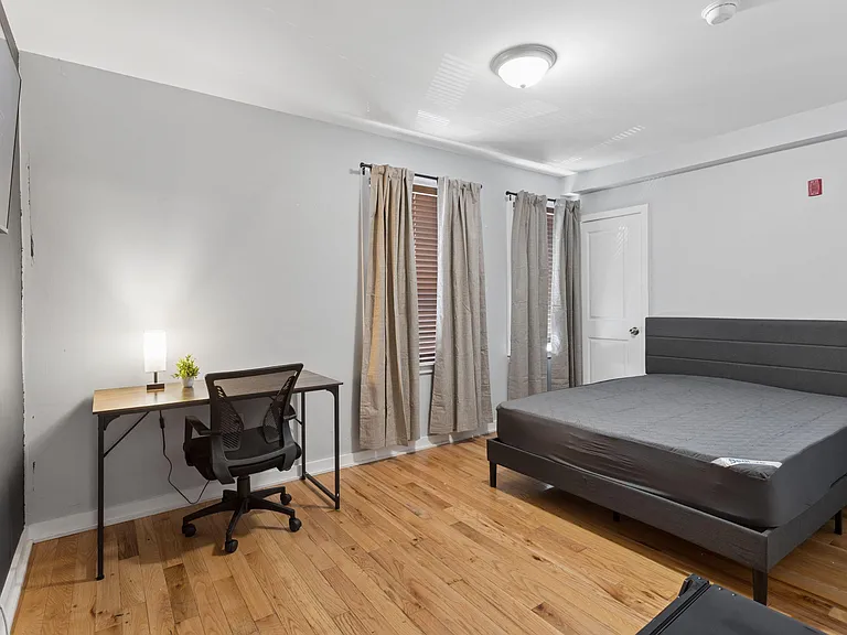 Furnished bedroom with hardwood floors, two windows with blinds and curtains, and a dedicated desk workspace.