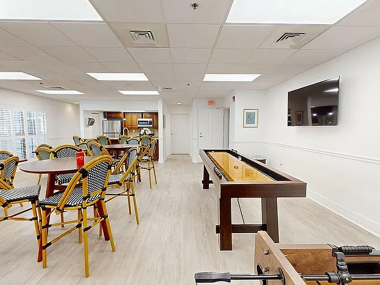 Inviting community room featuring stylish wicker chairs, large entertainment TV, and a shuffleboard table, perfect for social gatherings.