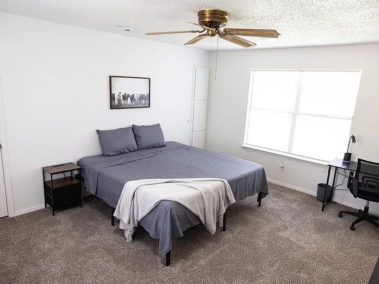 Furnished bedroom with a full bed, nightstand, and dedicated desk area, plus a large window and ceiling fan.